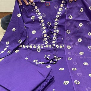 May include: A purple two-piece outfit with a long-sleeved top and pants. The top features a decorative neckline with a drawstring and is adorned with white circular patterns. The matching pants are folded neatly, and a sheer scarf is draped over the outfit.