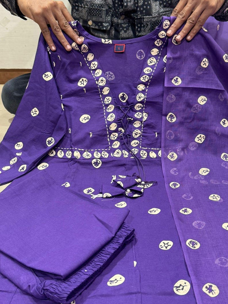 May include: A purple two-piece outfit with a long-sleeved top and pants. The top features a decorative neckline with a drawstring and is adorned with white circular patterns. The matching pants are folded neatly, and a sheer scarf is draped over the outfit.