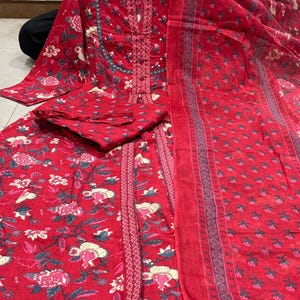 Może przedstawiać: Czerwony zestaw salwar kameez z kwiatowym nadrukiem. Zestaw zawiera tunikę z długim rękawem, pasujące spodnie i szalik. Tkanina ma powtarzający się wz&oacute;r r&oacute;żowych i białych kwiat&oacute;w, zielonych liści i niebieskich akcent&oacute;w. Tunika ma szczeg&oacute;łowy dekolt.