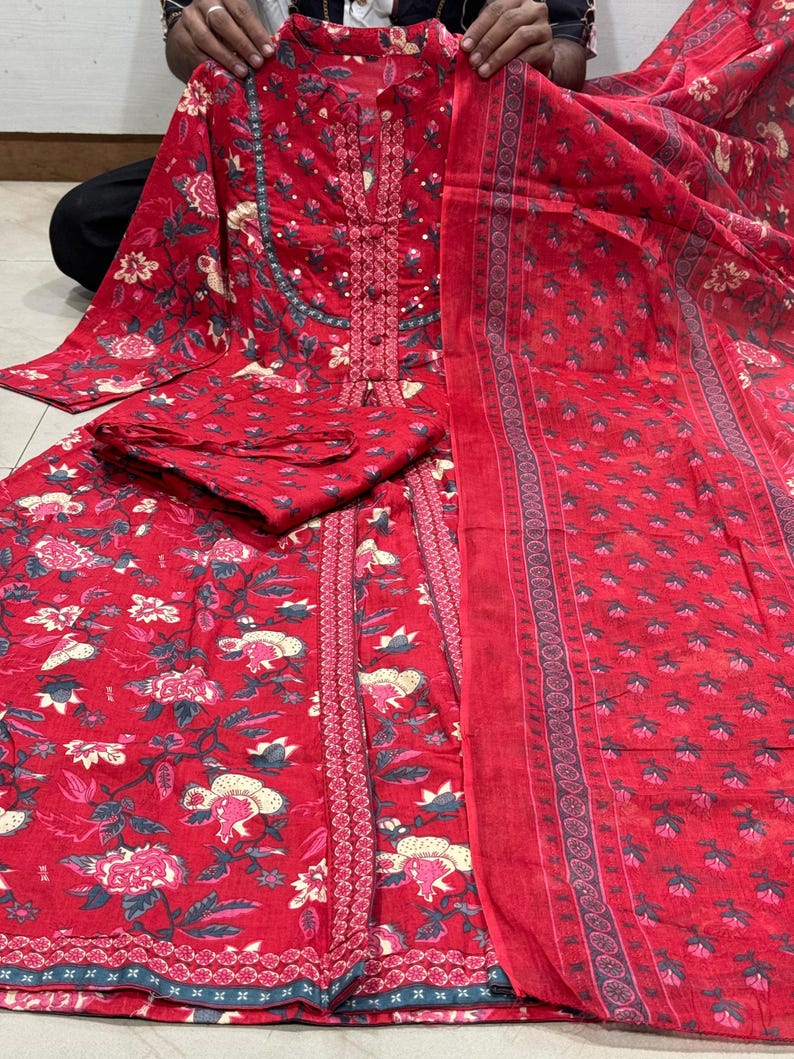 Może przedstawiać: Czerwony zestaw salwar kameez z kwiatowym nadrukiem. Zestaw zawiera tunikę z długim rękawem, pasujące spodnie i szalik. Tkanina ma powtarzający się wz&oacute;r r&oacute;żowych i białych kwiat&oacute;w, zielonych liści i niebieskich akcent&oacute;w. Tunika ma szczeg&oacute;łowy dekolt.