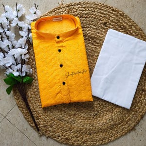 May include: A bright yellow kurta shirt with a textured pattern and black buttons, paired with white pants. The shirt is folded neatly on a woven mat, with white flowers and greenery to the left.