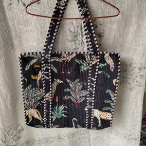 Pode incluir: Bolsa tote preta com estampa tropical com zebras, leopardos e flamingos em meio a folhagens exuberantes. A bolsa tem al&ccedil;as e acabamentos listrados em preto e branco e est&aacute; pendurada em um cabide vermelho.