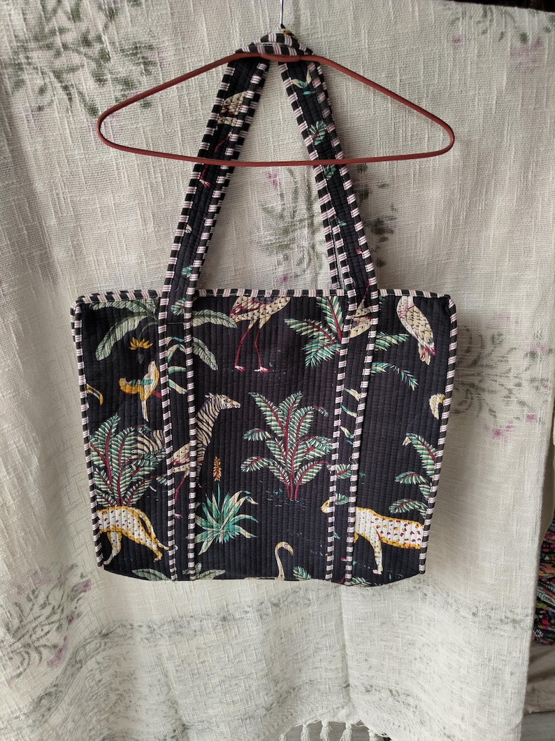 Pode incluir: Bolsa tote preta com estampa tropical com zebras, leopardos e flamingos em meio a folhagens exuberantes. A bolsa tem al&ccedil;as e acabamentos listrados em preto e branco e est&aacute; pendurada em um cabide vermelho.