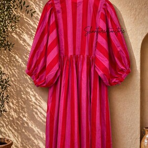 May include: A striking, striped dress in red and pink hangs on a wooden hanger. The dress has vertical stripes, a gathered waist, and voluminous sleeves. The text "Shantiercreationstore" is visible on the dress.