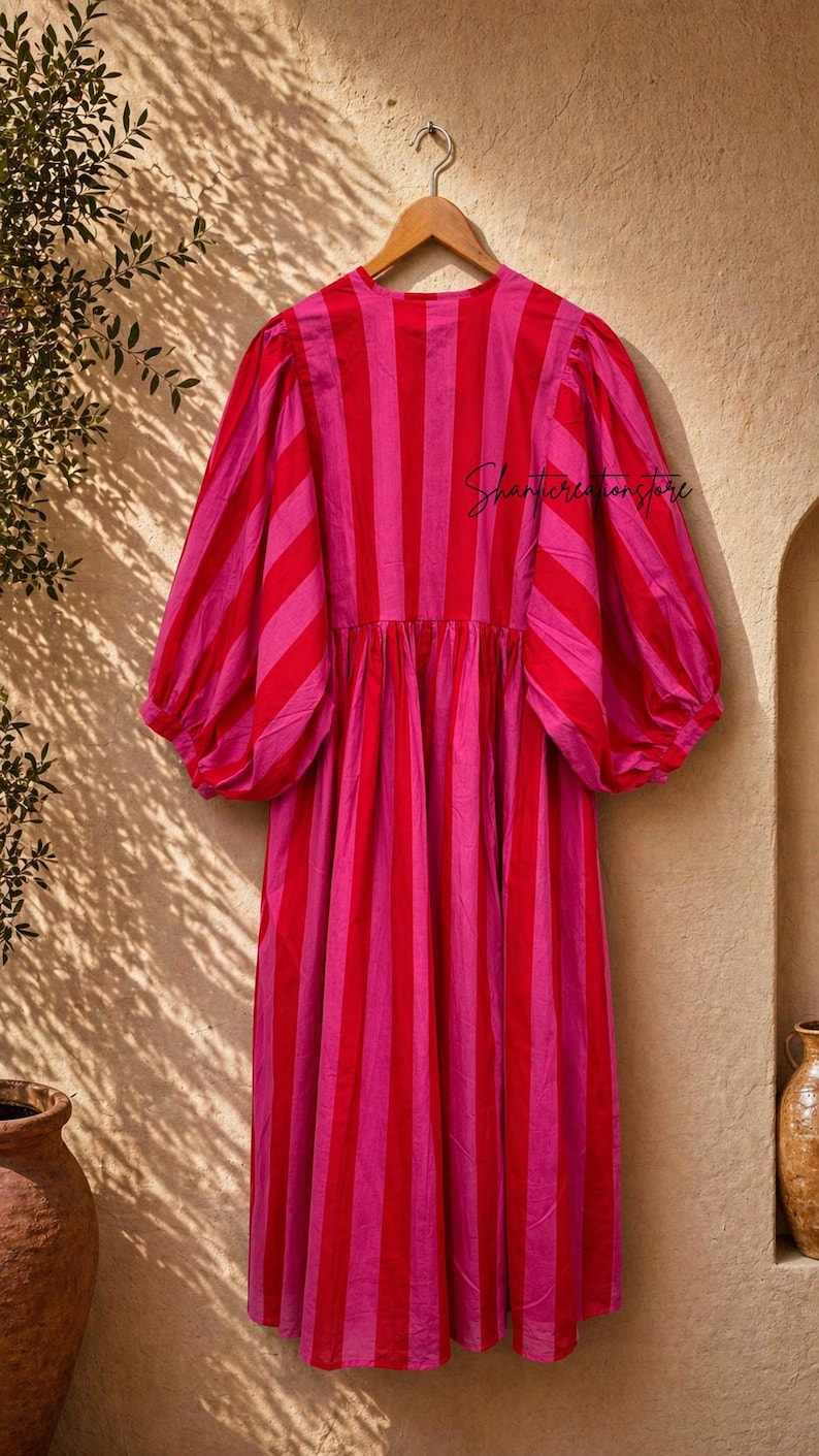 May include: A striking, striped dress in red and pink hangs on a wooden hanger. The dress has vertical stripes, a gathered waist, and voluminous sleeves. The text "Shantiercreationstore" is visible on the dress.