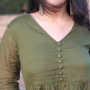 Peut inclure: Haut vert olive &agrave; manches longues avec un col en V et une rang&eacute;e de boutons assortis au centre. Le haut pr&eacute;sente un design fronc&eacute; et pliss&eacute; &agrave; la taille et est fait d'un tissu l&eacute;ger. Les manches sont longues et l&eacute;g&egrave;rement transparentes.