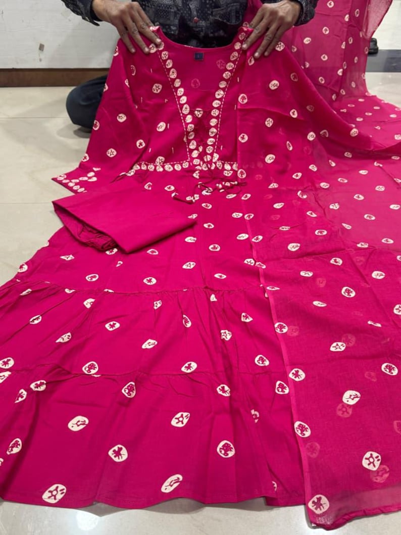 May include: A vibrant fuchsia-colored dress with a matching pair of pants. The dress features a tiered skirt and is adorned with white, circular patterns. The neckline is embellished with white embroidery. The outfit is made of a lightweight fabric.