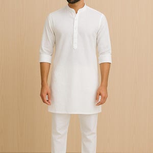 Pu&ograve; includere: Un completo kurta bianco e pantaloni abbinati. La kurta a maniche lunghe ha un colletto alla coreana e una chiusura frontale con bottoni. I pantaloni sono a gamba dritta. La persona indossa sandali marroni. Lo sfondo &egrave; un motivo a venature di legno chiaro.