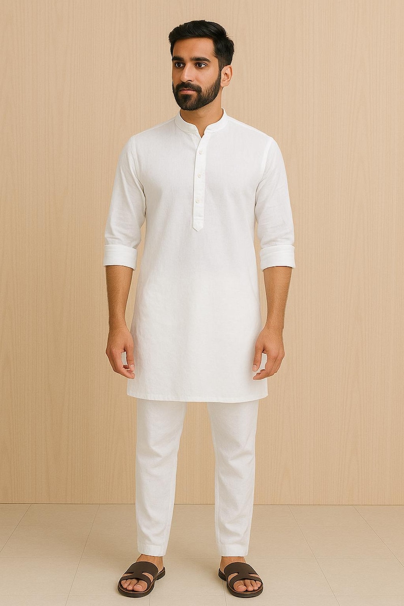 Pu&ograve; includere: Un completo kurta bianco e pantaloni abbinati. La kurta a maniche lunghe ha un colletto alla coreana e una chiusura frontale con bottoni. I pantaloni sono a gamba dritta. La persona indossa sandali marroni. Lo sfondo &egrave; un motivo a venature di legno chiaro.