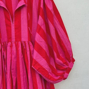 May include: A striking, striped dress in red and pink. The dress has vertical stripes, a gathered waist, and voluminous sleeves. The garment is hanging on a wooden hanger against a neutral background.