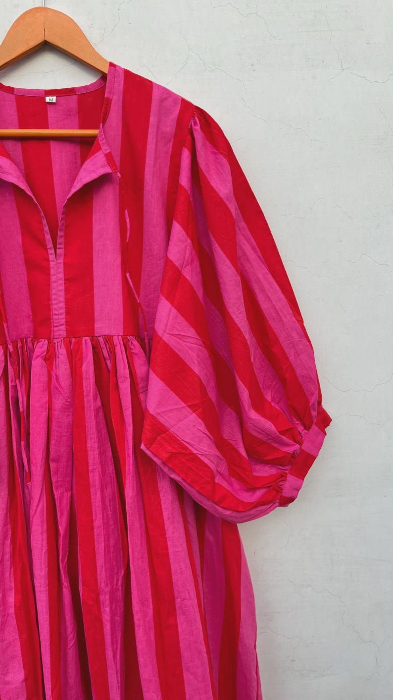 May include: A striking, striped dress in red and pink. The dress has vertical stripes, a gathered waist, and voluminous sleeves. The garment is hanging on a wooden hanger against a neutral background.
