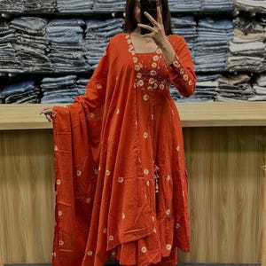 May include: An orange, long-sleeved dress with white circular patterns. The dress features a square neckline and a tiered skirt. A matching scarf is draped over the arm. The outfit is paired with matching pants and sandals.