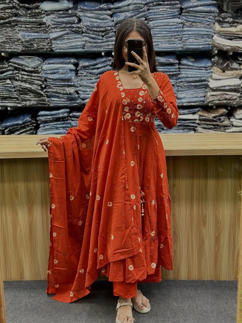 May include: An orange, long-sleeved dress with white circular patterns. The dress features a square neckline and a tiered skirt. A matching scarf is draped over the arm. The outfit is paired with matching pants and sandals.