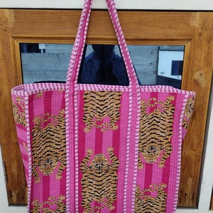 May include: A large pink tote bag with a tiger print design. The bag features a repeating pattern of tigers with black and brown stripes on a pink background with vertical pink stripes. The bag has long pink handles.