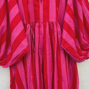 May include: A striking, striped dress in red and pink hues. The dress showcases broad vertical stripes, a V-neck, and voluminous sleeves. The fabric seems lightweight, ideal for warmer weather. The dress has a relaxed fit, with a gathered waist.