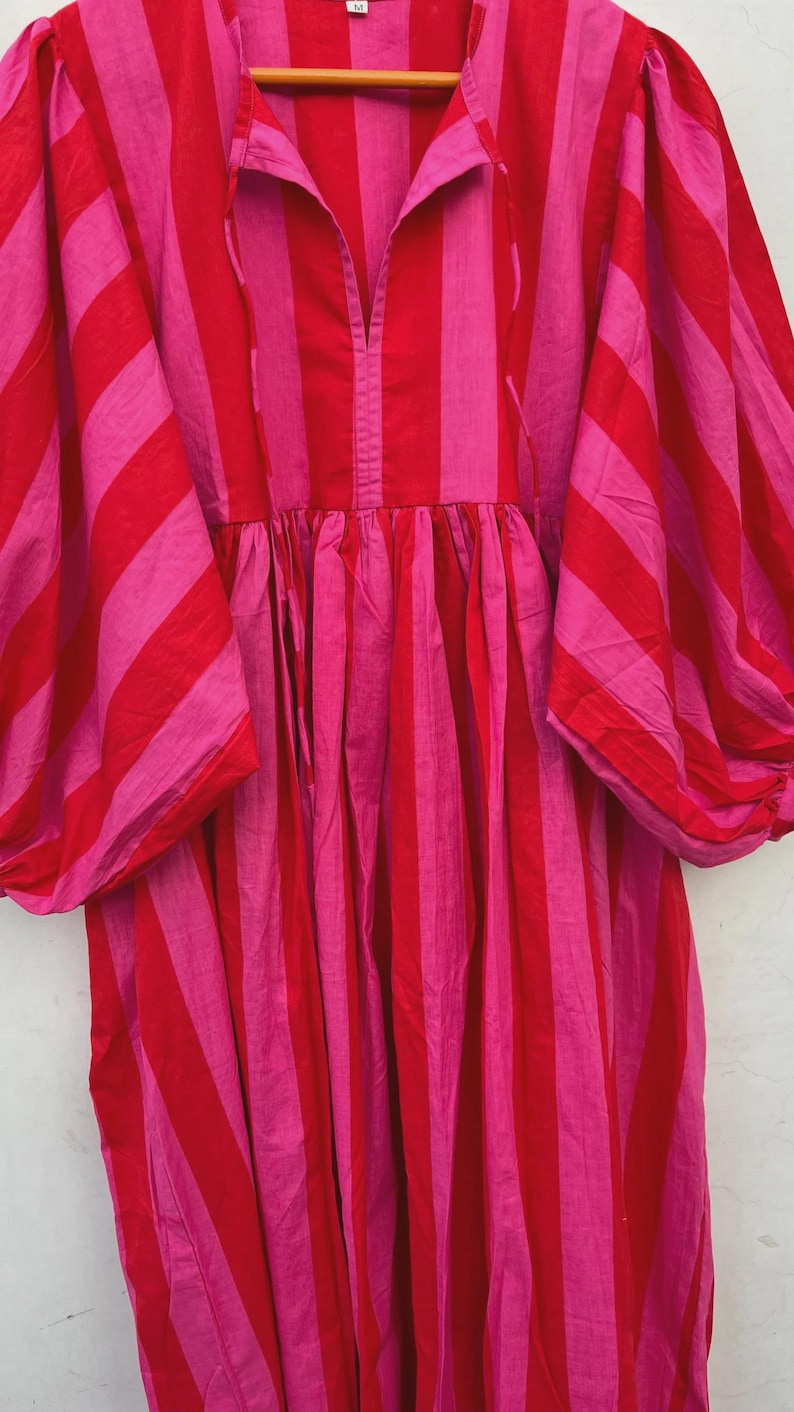 May include: A striking, striped dress in red and pink hues. The dress showcases broad vertical stripes, a V-neck, and voluminous sleeves. The fabric seems lightweight, ideal for warmer weather. The dress has a relaxed fit, with a gathered waist.