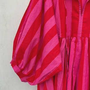 May include: A red and pink striped dress with a gathered waist and voluminous sleeves. The dress has a collared neckline and a button-down front. The fabric appears to be lightweight and suitable for warm weather.
