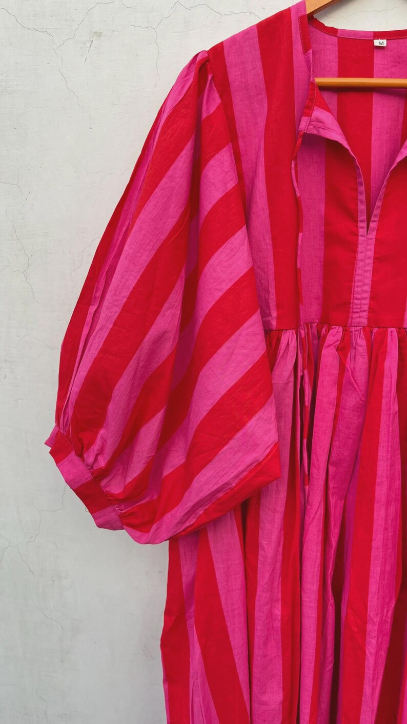 May include: A red and pink striped dress with a gathered waist and voluminous sleeves. The dress has a collared neckline and a button-down front. The fabric appears to be lightweight and suitable for warm weather.