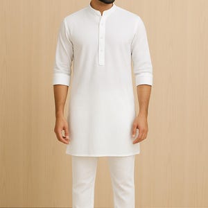 Pu&ograve; includere: Un completo tunica e pantaloni bianchi. La tunica ha un colletto alla coreana e una paramontura abbottonata. I pantaloni sono a gamba dritta. La persona indossa sandali in pelle marrone. L'outfit &egrave; uno stile tradizionale, spesso indossato in Asia meridionale.