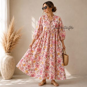 May include: A long, flowing dress with a floral pattern in shades of pink, yellow, and purple. The dress has a V-neckline with a tie detail and three-quarter sleeves. A woven straw handbag is held in the hand.