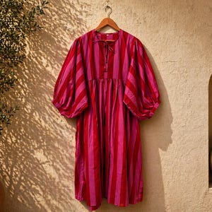 May include: A red and pink striped dress with short, puffed sleeves hangs on a wooden hanger. The dress has a loose, flowing shape and a tie detail at the neckline. The background is a textured beige wall with shadows.