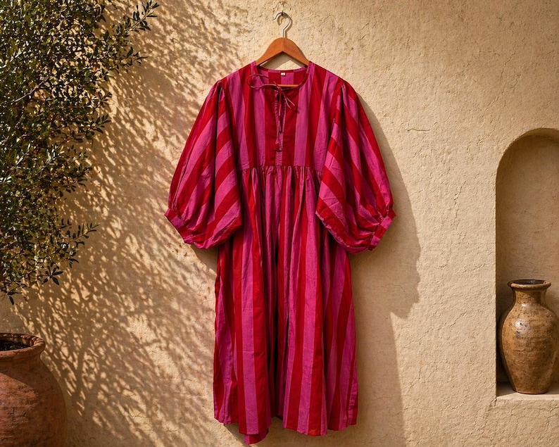 May include: A red and pink striped dress with short, puffed sleeves hangs on a wooden hanger. The dress has a loose, flowing shape and a tie detail at the neckline. The background is a textured beige wall with shadows.