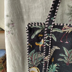 Pode incluir: Bolsa tote preta com um design acolchoado com uma cena da selva com zebras, p&aacute;ssaros e palmeiras. A bolsa tem al&ccedil;as e acabamentos listrados em preto e branco com detalhes em rosa. A bolsa &eacute; feita de um material macio e acolchoado.