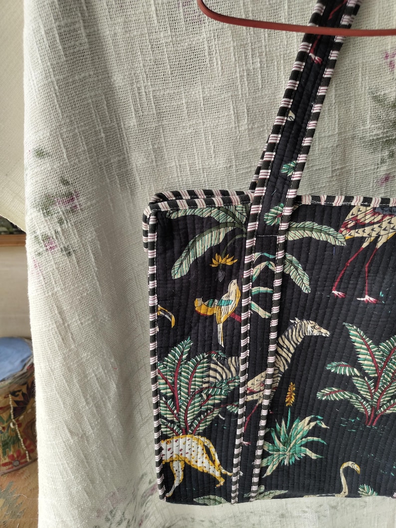 Pode incluir: Bolsa tote preta com um design acolchoado com uma cena da selva com zebras, p&aacute;ssaros e palmeiras. A bolsa tem al&ccedil;as e acabamentos listrados em preto e branco com detalhes em rosa. A bolsa &eacute; feita de um material macio e acolchoado.