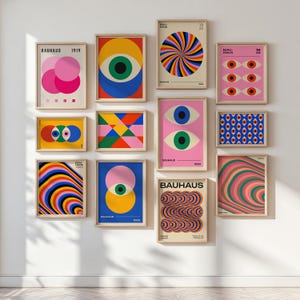 May include: A collection of framed Bauhaus art prints. The prints feature geometric shapes, eye motifs, and abstract designs in vibrant colors like pink, blue, yellow, and green. Each print is in a wooden frame, creating a gallery wall aesthetic.