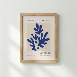 May include: A framed art print featuring a blue abstract design on a beige background. The print includes the text "PAPIERS DÉCOUPÉS" and "MATISSE PARIS, FRANCE 1955". The frame is made of light-colored wood.