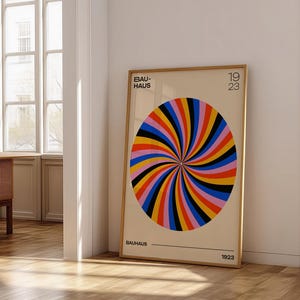 Puede incluir: Un póster Bauhaus enmarcado con un diseño en espiral colorido. La obra de arte presenta un patrón circular con segmentos rojos, naranjas, amarillos, negros, azules y rosas. El póster está en un marco de madera y se apoya en una pared blanca.