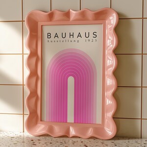 May include: A pink framed art print with the word "BAUHAUS" at the top and a pink and white arched design. The frame has a wavy, scalloped edge. The art print is displayed against a tiled background.
