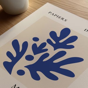 以下が含まれることがあります： ベージュの背景に青い抽象的な花の模様がプリントされたアートプリント。上部には黒で「PAPIERS」の文字が印刷されています。白い紙に印刷されています。白いマグカップが見えます。