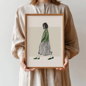 Puede incluir: Una impresión artística enmarcada con una ilustración en acuarela de una persona caminando. La figura lleva un suéter verde, una falda a rayas y zapatos verdes. La obra de arte tiene una paleta de colores neutros y un marco de madera.