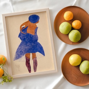 May include: A framed watercolor painting of a figure in a blue dress and hat, with brown skin and purple boots. Two wooden trays hold oranges and green fruit. The painting is in a light wood frame.