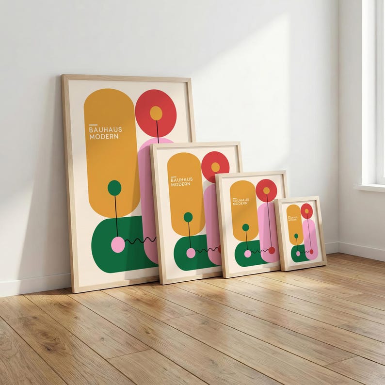 May include: Four framed Bauhaus Modern art prints in varying sizes lean against a white wall. Each print features a geometric design with mustard yellow, pink, green, and red shapes. The prints are displayed on a wooden floor.
