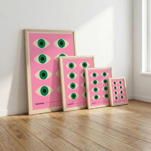 May include: Four framed art prints featuring a repeating eye design on a pink background. The eyes are green with off-white eyelids. The word "BAUHAUS" is printed at the bottom of the largest print. The frames are light wood.