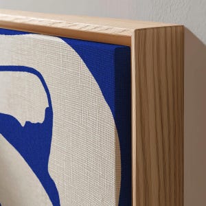 Puede incluir: Impresión en lienzo de arte abstracto con un diseño audaz en azul y blanco roto. La obra de arte está estirada sobre un marco de madera, mostrando una estética moderna. El lienzo tiene una apariencia texturizada, agregando profundidad. El marco de madera tiene una veta natural.