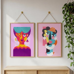 Puede incluir: Dos impresiones artísticas abstractas enmarcadas. La obra presenta rostros geométricos coloridos con líneas y formas audaces. Las impresiones están en marcos dorados y cuelgan de una pared blanca. Una planta cuelga a la derecha.