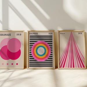 May include: Three framed Bauhaus art prints. The prints feature geometric designs in pink, black, and rainbow colours. Each print is labelled "BAUHAUS 1919". The frames are a light wood colour, and the background is a neutral beige.