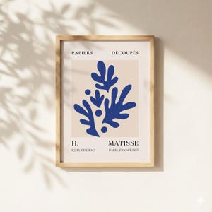 Könnte beinhalten: Ein gerahmter Kunstdruck mit einem blauen abstrakten Design auf beigem Hintergrund. Der Druck enthält den Text „PAPIERS DÉCOUPÉS“ oben und „MATISSE PARIS, FRANCE 1955“ unten. Der Rahmen ist aus hellem Holz.