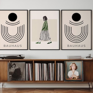 Może przedstawiać: Trzy oprawione grafiki Bauhaus z czarnymi ramami i nowoczesna szafka na płyty z połowy wieku. Grafiki przedstawiają geometryczne wzory i postać. Szafka jest wykonana z drewna i mieści płyty winylowe i gramofon. Na grafikach wydrukowano słowo "BAUHAUS".
