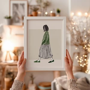 May include: A framed art print showing a watercolour illustration of a person walking. The figure is wearing a green sweater, a striped skirt, and green shoes. The artwork is set against a neutral background, framed in a white border.