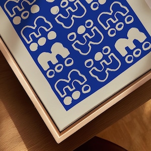 May include: A framed canvas art piece featuring a repeating pattern of abstract shapes in white against a vibrant blue background. The artwork is set within a natural wood frame, creating a modern aesthetic. The canvas is displayed on a wooden surface.