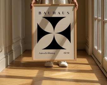 May include: A framed Bauhaus poster with a geometric black and white design. The poster features a black cross-like shape on a cream background, with the word "BAUHAUS" at the top and "Ausstellung 1919" at the bottom. The frame is light brown.
