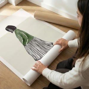 以下が含まれることがあります： 緑色のジャケットとストライプのスカートを着た人物の水彩イラストが描かれたアートプリント。白いチューブからプリントが広げられています。アートワークは、大きなオフホワイトの紙にあります。
