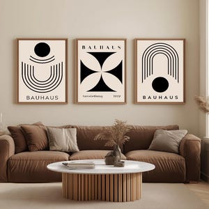 Può includere: Tre stampe artistiche Bauhaus incorniciate con disegni geometrici in tonalità nere e beige. Le stampe sono esposte sopra un divano marrone, creando un'estetica moderna e minimalista. La parola "Bauhaus" è stampata su ogni cornice.
