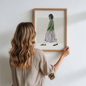 May include: A framed art print showing a watercolour illustration of a person walking. The figure is wearing a green jacket, a striped skirt, and green shoes. The artwork is in a wooden frame and is held against a white wall.