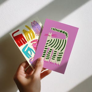 Könnte beinhalten: Eine Hand hält drei farbenfrohe Katzenkunstdrucke. Ein Druck ist lavendelfarben mit einer grün-weiß gestreiften Katze. Der andere Druck hat einen weißen Hintergrund mit roten, gelben, blauen und rosa Katzenillustrationen. Die Drucke sind rechteckig.