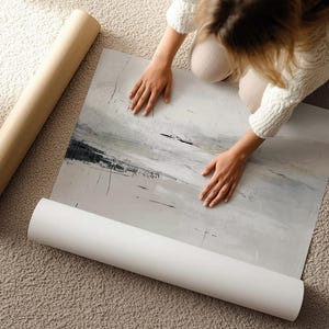 Puede incluir: Una impresión de arte abstracto en tonos grises, blancos y negros se desenrolla sobre una alfombra. También se ven un rollo de impresión y un tubo de papel marrón. La obra presenta pinceladas y un diseño similar a un paisaje.